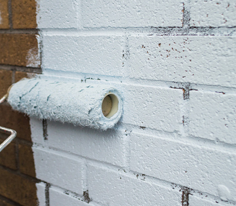 person painting a wall with a roller tree in background three coats of paint